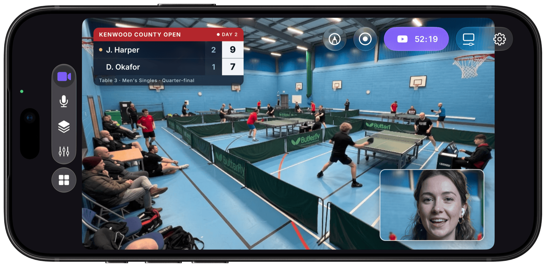 Camo Studio on iPhone in landscape, live on YouTube from a local table tennis tournament: a wide shot of the sports hall with a Kenwood County Open score bug in the corner and a commentator picture-in-picture