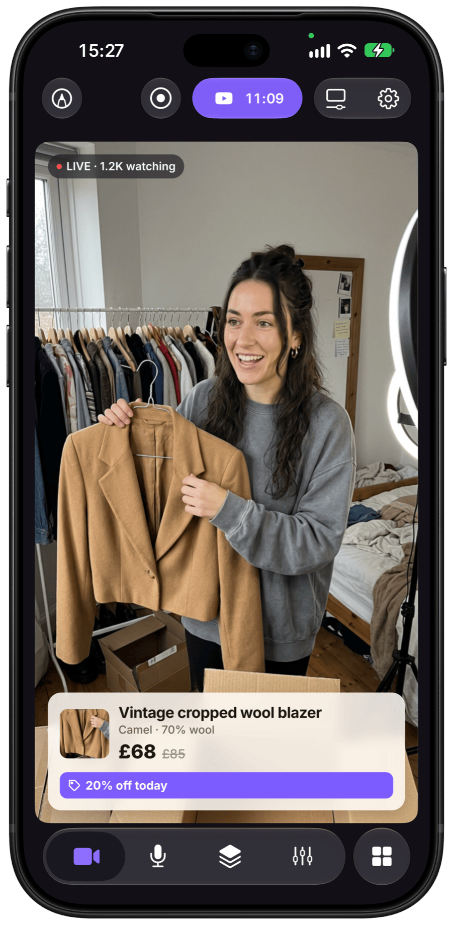 Camo Studio on iPhone in portrait, live on YouTube: a vintage reseller in her spare room, surrounded by a clothing rail and shipping boxes, holds up a camel cropped wool blazer, with a product card showing the price of £68 and 20% off today